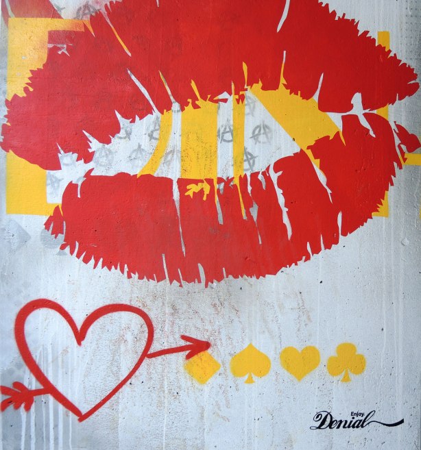 picture of art (mural) on a concrete support holding up a road above a skateboard and basketball park - bottom part of a pillar with a painting of a big red set of lips, a red heart with an arrow through is as well as yellow symbols from playing card - hearts, diamond, spage and club.  Signed Enjoy denial. 