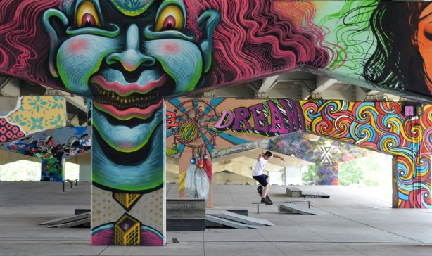 picture of art (mural) on a concrete support holding up a road above a skateboard and basketball park - young man on skateboard jump in the park with many of the pillars in the photo including a large greyish blue clown face 