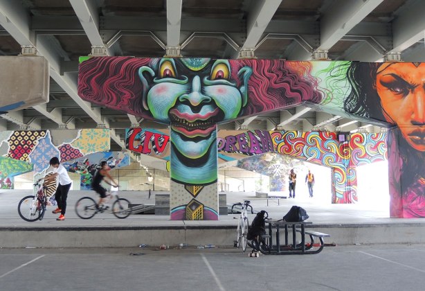 picture of art (mural) on a concrete support holding up a road above a skateboard and basketball park - guys on bikes in the park with many of the pillars in the photo including a large greyish blue clown face and an orange woman's face. 