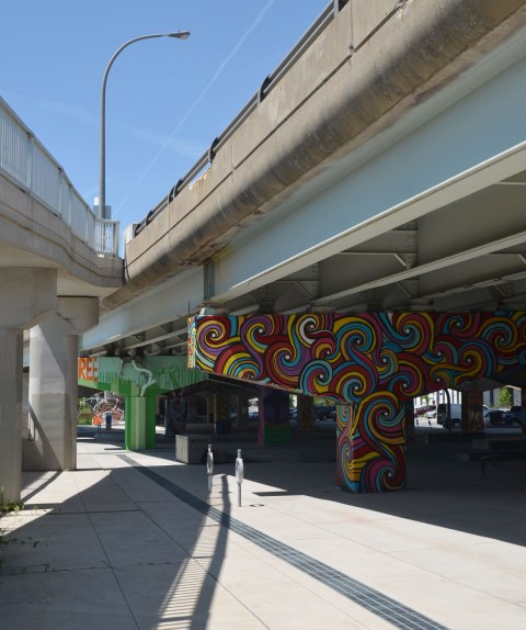 Two overpasses come together to form one, view from below, there is an urban park under the overpass on the right, with the concrete supports holding up the road covered with paintings by street artists.