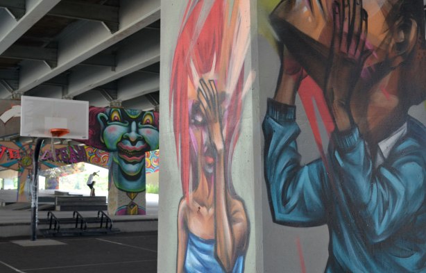 picture of art (mural) on a concrete support holding up a road above a skateboard and basketball park -