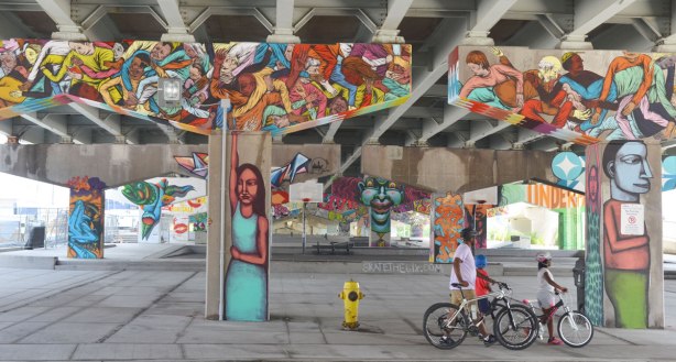 A man and two kids walk their bikes past Underpass Park on Lower River St. in Toronto.  A road is above them.  Concrete supports that hold up that road have been decorated with paintings by various street artists.  Closest to the street, the pillars are people with their arms raised so it looks like they are holding up the road.  The horizontal part of the supprts are covered with pictures of people flying outward from the center. 