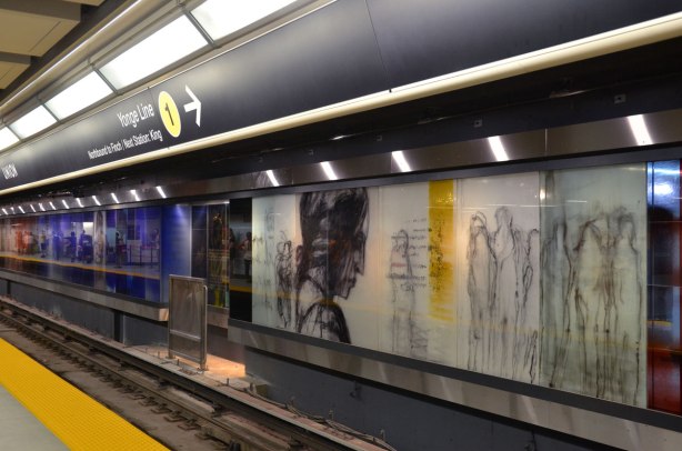 Looking along a subway platform at Union Station, the far wall is an art installation, paintings on glass panels of people 
