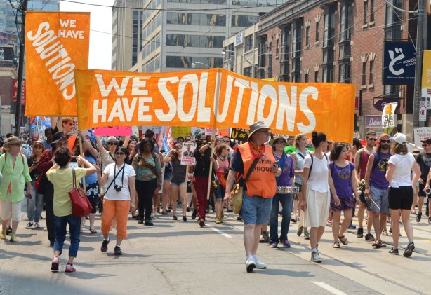 Marchers on the jobs, justice and climate action protest march who are carrying a large orange banner that says We have solutions. 