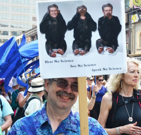 A man is holding up a protest sign with Prime Minister Stephen Harper's head on three monkeys along with the worsd, Hear no science, speak no science, see no science. 