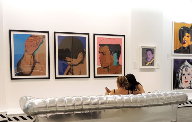Two women are sitting ona silver couch in an art gallery.  On the wall in front of them are 3 silkscreen Andy Warhol prints of Mohammad Ali.  