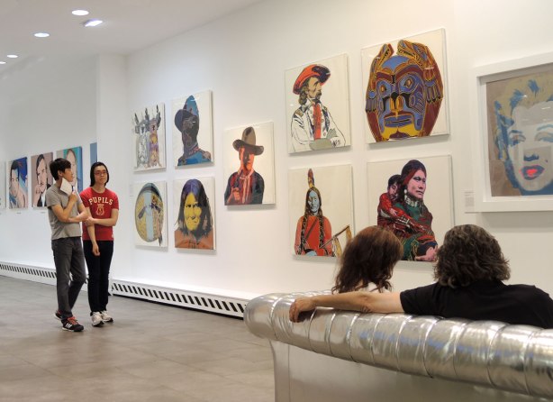 Some of Warhol's Cowboys and Indians series prints hang on a gallery wall as two young Asian men walk past. There is also a couple sitting on a silver coloured couch in the right side of the photo.