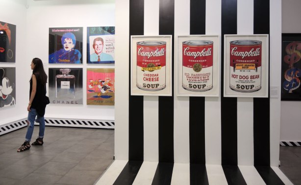 prints of three Campbells soup cans on a black and white striped wall, cheddar cheese, old fashioned vegetable, and hot dog bean.  To the left a woman is looking at other Andy Warhol prints on a wall including a print of Mickey Mouse, Lucille Ball and a young Ronald Regan. 