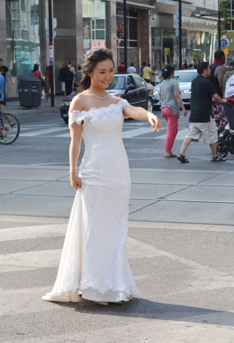 A bride is standing in the middle of an intersection in downtown Toronto, wearing a long white wedding dress, she has her arm up and is beckoning to the groom. 