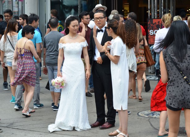 A bride and groom are standing on the corner of Yonge and Dundas streets in Toronto.  They are talking to a woman in a white dress who is organizing the wedding photo shoot. 