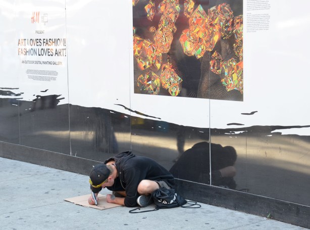 A man is sitting on the sidewalk. He is making a sign on a piece of brown cardboard. The wall behind him has a large picture on it that is part of an exhibit