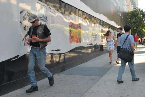 People walk by the hoardings beside the sidewalk on Dundas St. near Yonge, part of the new H & M store being built there. There are pictures on the wall.
