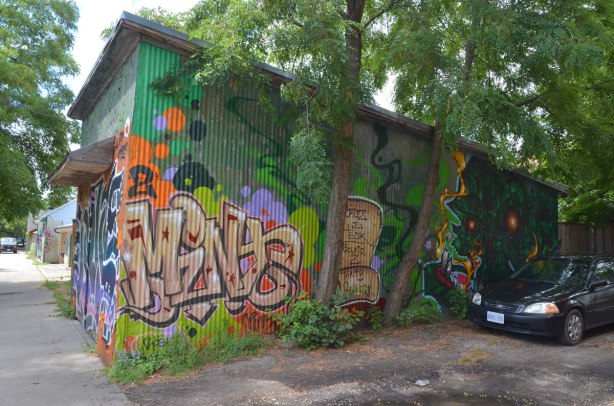 The side of a garage door in summer, two trees and a parked car.  The side of the garage is covered with street art 