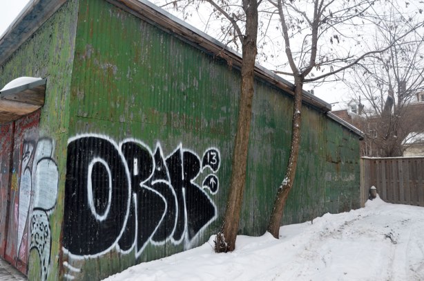 The side of a garage in an alley in winter, snow on the ground, two small trees.   The side of the garage is painted green and it has a black and white orak tag on it. 
