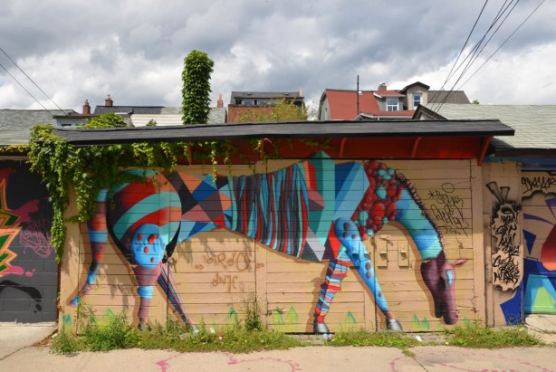 A painting by street artist birdo of a red and blue striped horse like creature with its neck bent downwards so that it looks like it is eating the weeds that are growing between the cracks in the lane.  It is on a garage door, tops of houses can be seen in the background, above the garage