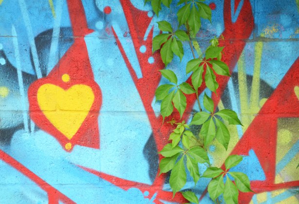 close up picture of a green leafed vine growing up a street art painting in blue and red with a yellow heart 