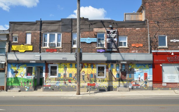 looking across the street to a row of brick store fronts, two storeys high. The bottom storey has been painted with a mural that has a yellow background.