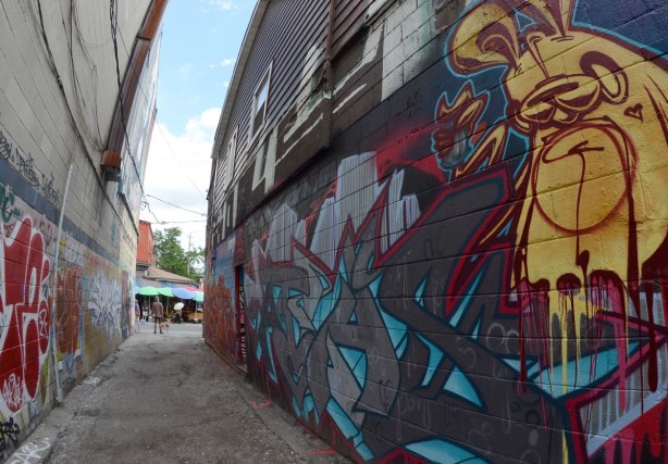 looking down the alley, wide angle shot, poser bunny mural on the right side, red tag graffiti on the left.  At the end of the alley a man stands with his dog, a woman with a white umbrella is passing by 