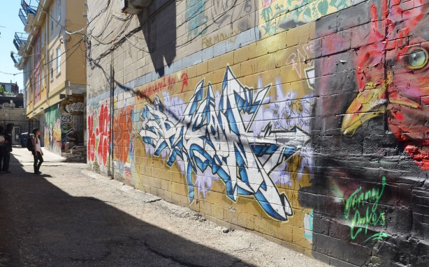 a wall in a lane, a large red chicken head painting is on the right and it seems to be looking down the lane.   A woman is standing in the lane 