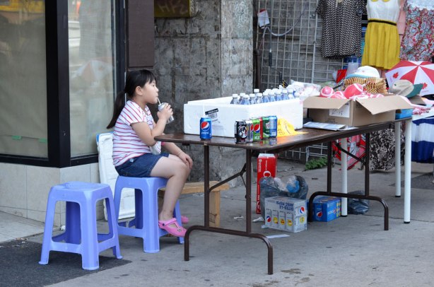 A girl tends a table full of cold drinks that are for sale, on the sidewalk