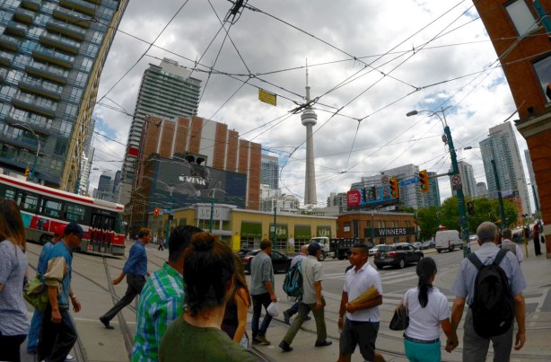 the intersection of King and Spadina in Toronto with a wide angle lens. People are crossing the street, there is a streetcar and lots of streetcar wires. THe LCBO and Winners are in the background as well as the CN Tower