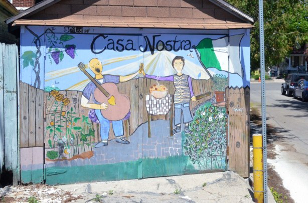 garage door with a picture on it titled Casa Nostra, a picture of a man playing a large guitar and a woman standing beside an outdoor table with food on it. 