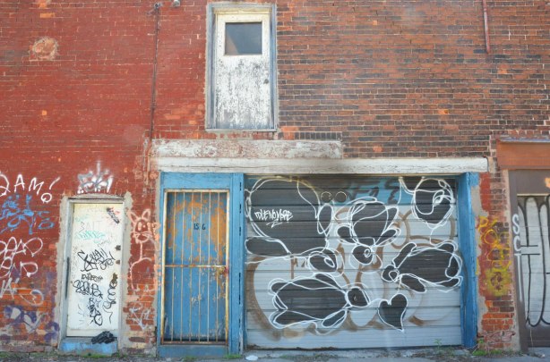 blue door with faded paint and a metal grille in front of it, beside a garage door with graffiti on it including the words Idle no more. Upper storey has an exterior door that goes nowhere 