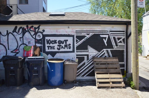 black and white street art on a garage door that is partially obscured by trash bins and wooden structure. The art is geometric shapes and includes the words Kick out the Jams