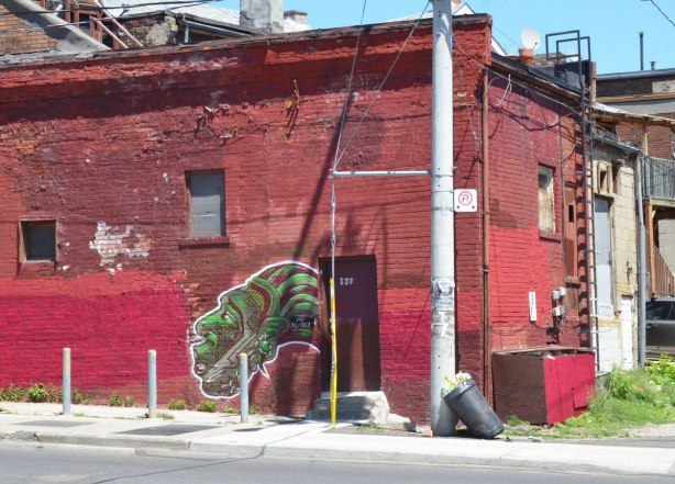 On a wall that has been painted a couple of shades of red is a painting of a green head with a green and red striped head dress on 