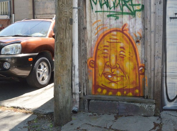 A brown car is parked in a driveway beside a door with a yellow man's face painted on the bottom half of the door. He's bald, little round ears and his eyes are closed. 