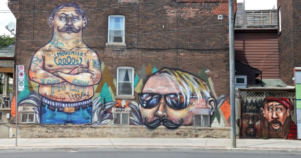 Movember mural on the side of a two storey brick house.  On the left is a man, standing, shirtless with just the top of his jeans showing.  He is tattoed.  Beside him is just the upper part of another man's face, from the large moustache to the top of head with thinning hair.  To the right are two small men's heads, both with moustaches, painted on a wooden fence behind the house. . 
