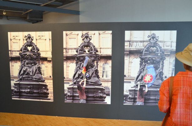 In an art gallery, a series of three large photographs of a statue of Queen Victoria.  THe first picture is just the statue, the middle picture is a man starting to put a  flag over the bottom part of the statue and the third picture is the flag on the statue.  Flag is solidarity flag created by Scott Benesiinaabandan, black and blue background, red circle in the middle, yellow sun in the red circle