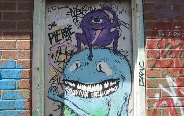 A door in an alley with two montsters painted on it. A one eyed purple monster with a long pink tongue is sitting on top of a blue monster who is trying to pinch the end of the purple monster's tongue