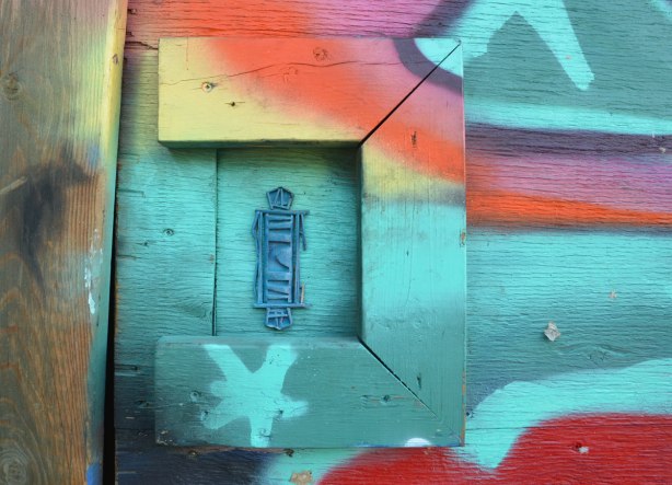Close up of a graffiti piece, many colours, with a blue 3D stick figure surrounded on three sides by frame-like boards