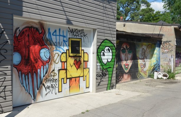 Two garage doors with street art. On the left is a yellow lovebot and a red grominator. On the second is a large woman's face that has been scribbled on as well as a fairy-like creature. The latter two are the work of EGR.An old broken toilet is in the corner. 