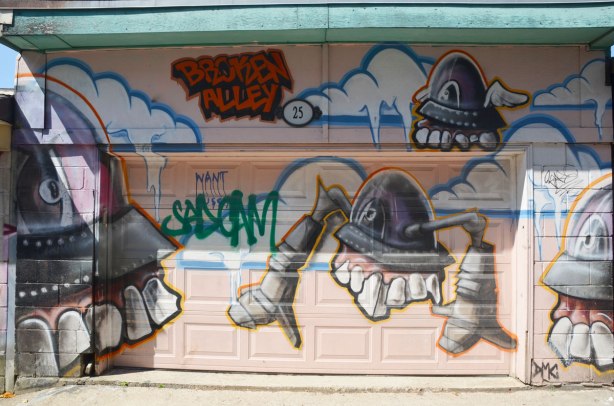 A mural of big headed, big toothed creatures by broken alley on a garage door in a lane. 