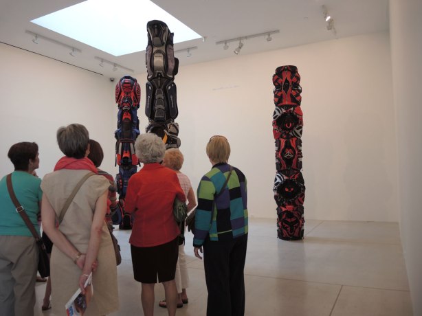 A group of women looks at an art installation of three large totem poles made of golf bags on display in an art gallery (Art Gallery of Ontario)