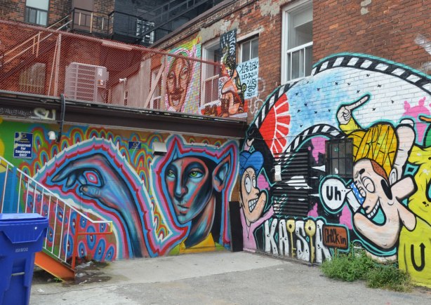 Two walls covered with street art. One wall has a mural for the Kaisar guest house. The other wall has a man's head and hand that is pointing to an exterior staircase. 