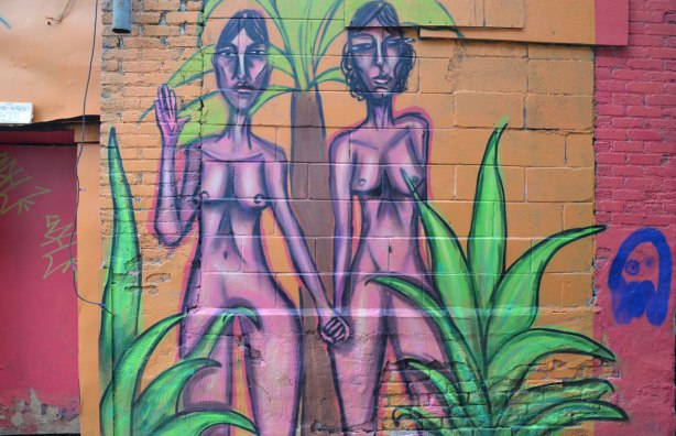 mural street art painting of a naked woman, two pink women holding hands, drawn with purple outlines, on a pink background, with some green plants in the painting too 