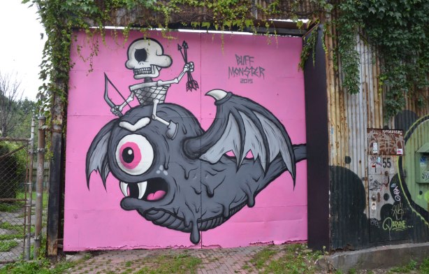 A garage door in an alley painted pink. A large fat grey creature with bat-like wings and bulging eye takes up most of the space, a small skeleton is sitting on the creature's head. The piece is signed Buff Monster 
