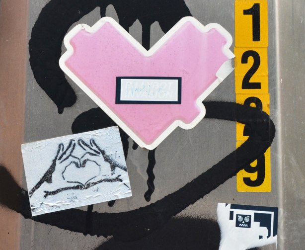 Two heart stickers on a metal pole. One is a pink lovebot shaped heart and one is a drawing of two hands making a heart shape with the thumbs and forefingers