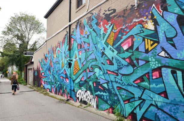 A large street art piece painted in an alley. It covers most of the side of a brick house. It is mostly blue lines and arrows. 