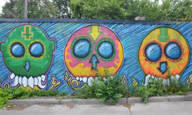 3 big round headed aliens by street artist mska, one is green, one is pink and yellow and one is yellow and orange, on a fence in an alley 