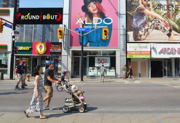 A couple is pushing a stroller up Yonge St. past some of the stores. No cars on the street 