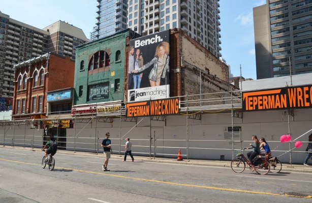 A few people walk on the streets, two bikes also, the bikes have pink balloons attached to them, they are on Yonge Street, just south of Bloor in front of the construction on the west side of Yonge, the lower storey of old brick stores is boarded up, the upper storeys are still visible