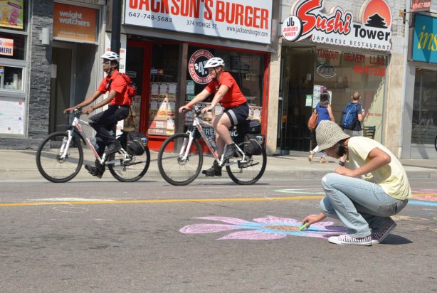 Two men wearing red T shirts are riding their bikes on Yonge street, passing a woman who is using chalk to draw a floor on the street