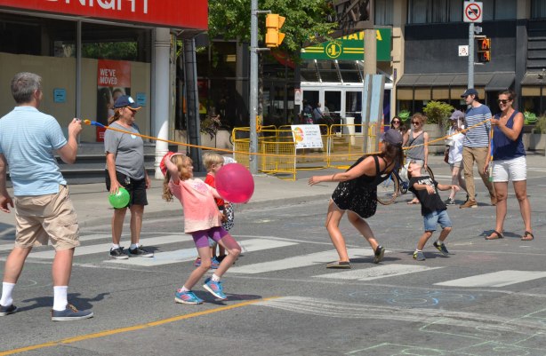 A group of adults and kids are doing the limbo with a long rope on the street on a day when the road is closed to traffic. 