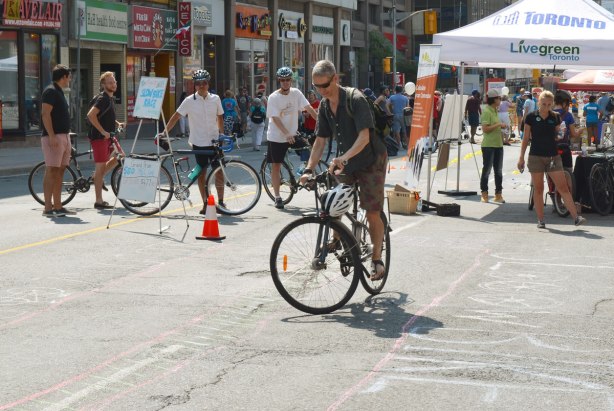 A man is riding his bike as slowly as possible and still stay within a narrow path as part of a slow bike race. Other people are standing beside their bikes and watching him