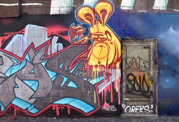 a yellow poser bunny on a wall in an alley beside a door that is covered with tags including oreks 