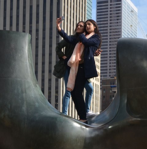 People taking selfies - two young women are on top of The Archer sculpture in Nathan Phillips Square. Downtown skyscrapers are in the background 